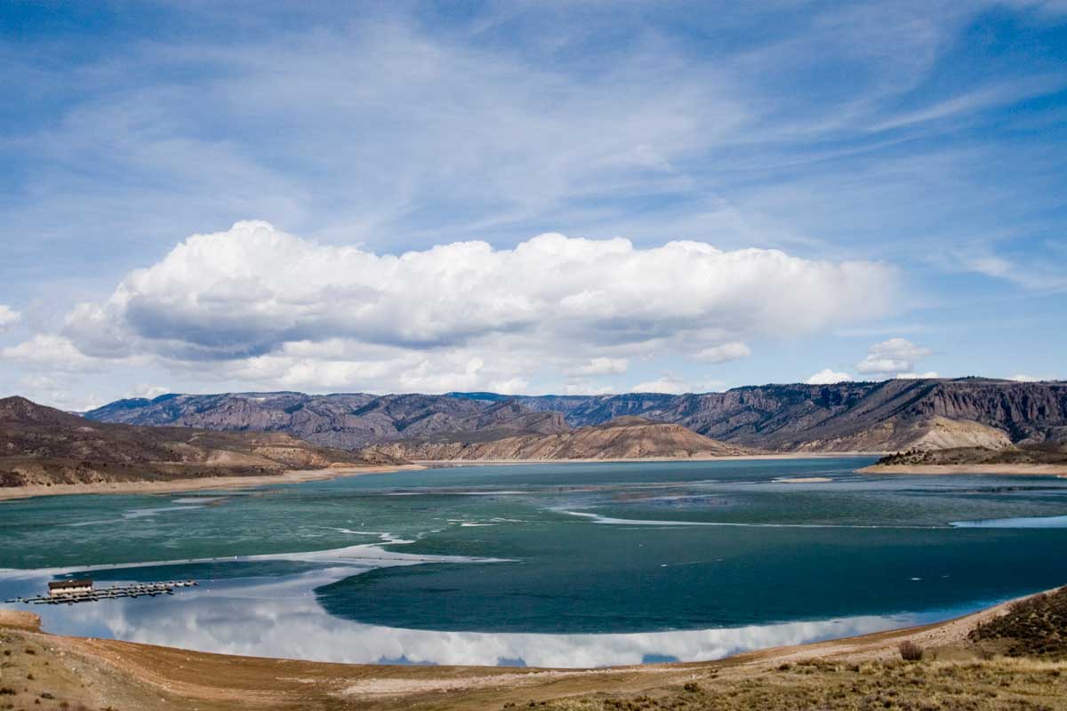 The sandy shores of Blue Mesa Reservoir look onto the blue water with a rocky peaks in the background with a blue sky and white clouds.
