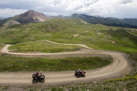 Two ATVers zoom down a winding dirt road split with green-grass fields. In the distance are jagged peaks under a cloudy sky in the San Juan Mountains.