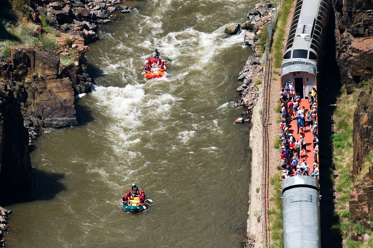 Two groups of whitewater rafters paddle down a swift river near the Royal Gorge in Colorado. On shore, visitors watch from an open section of what looks like a subway car train.