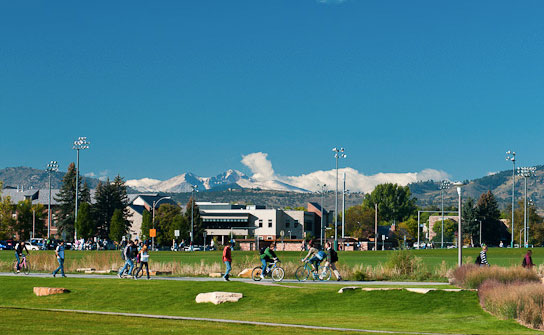Students bike and walk on a Colorado State University campus path. In the background are university buildings and snow-covered peaks.