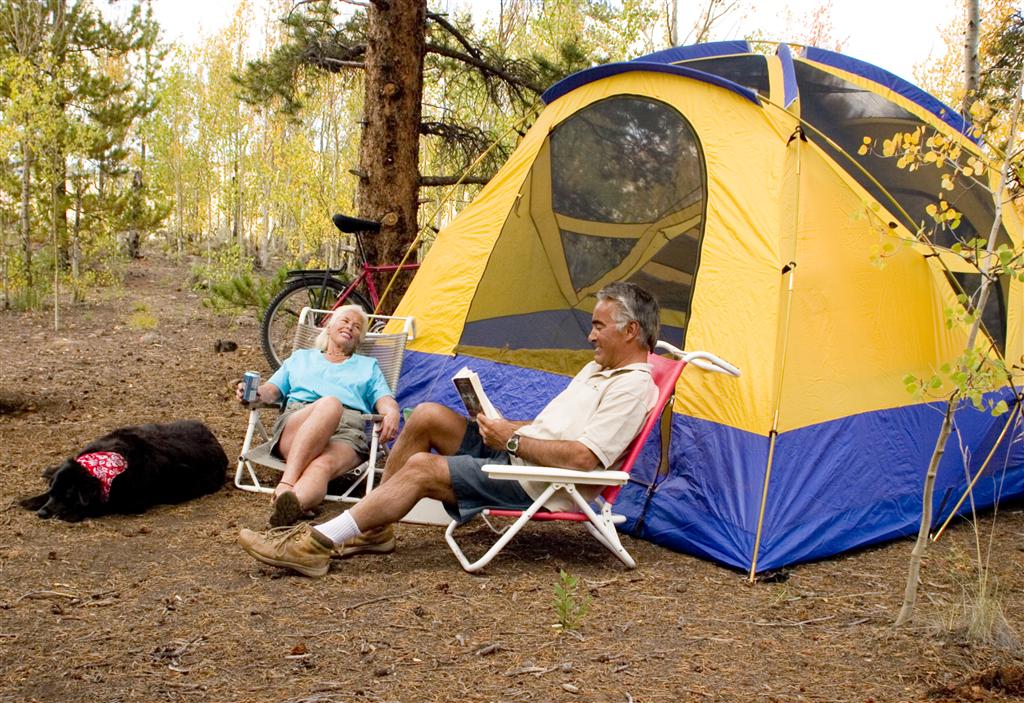 A man and woman sit in camp chairs in front of a yellow and blue tent in Crawford State Park. There's a dog sleeping to the left of the woman, who is wearing a blue shirt. There are skinny green trees in the background.