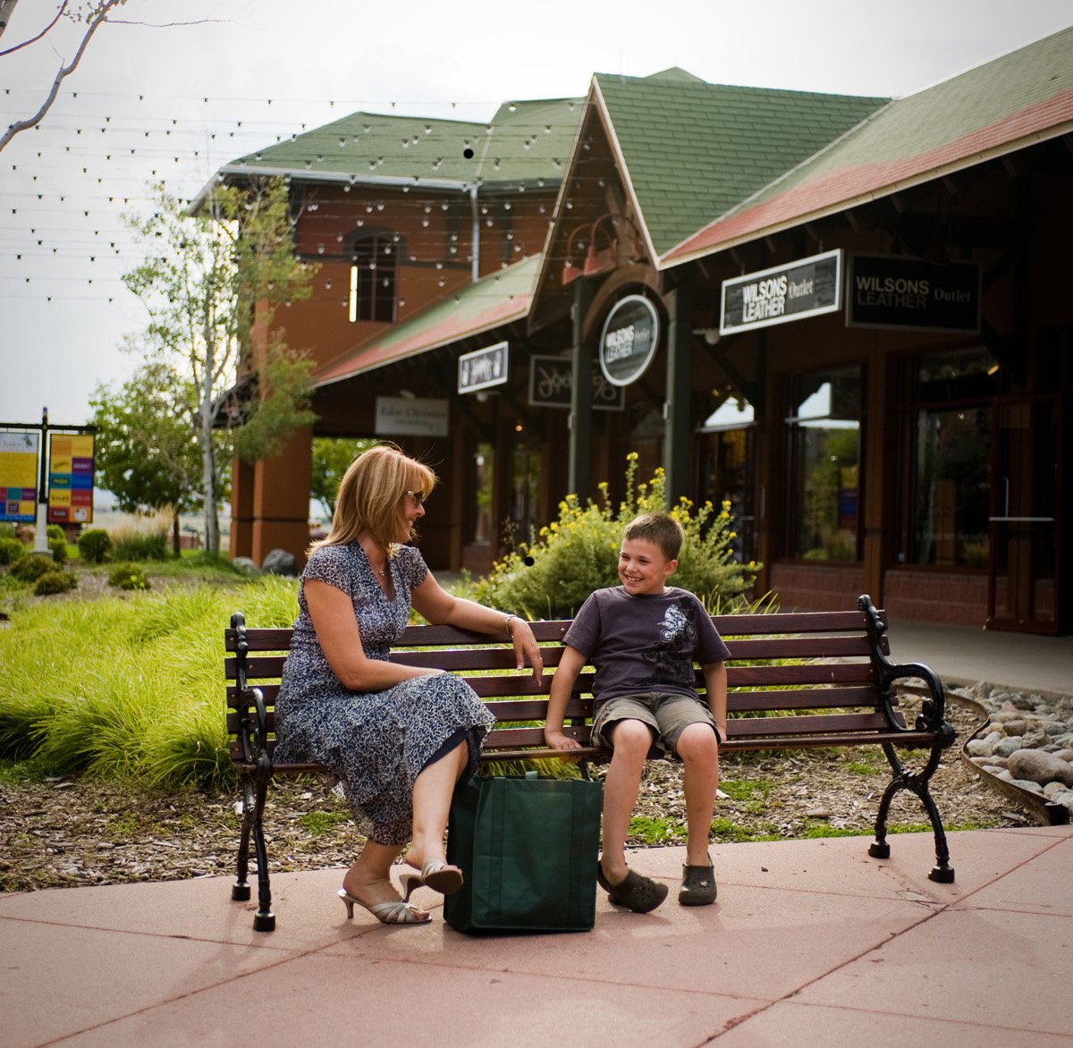 A woman sits on a bench with a child at an outlet mall in the summer.
