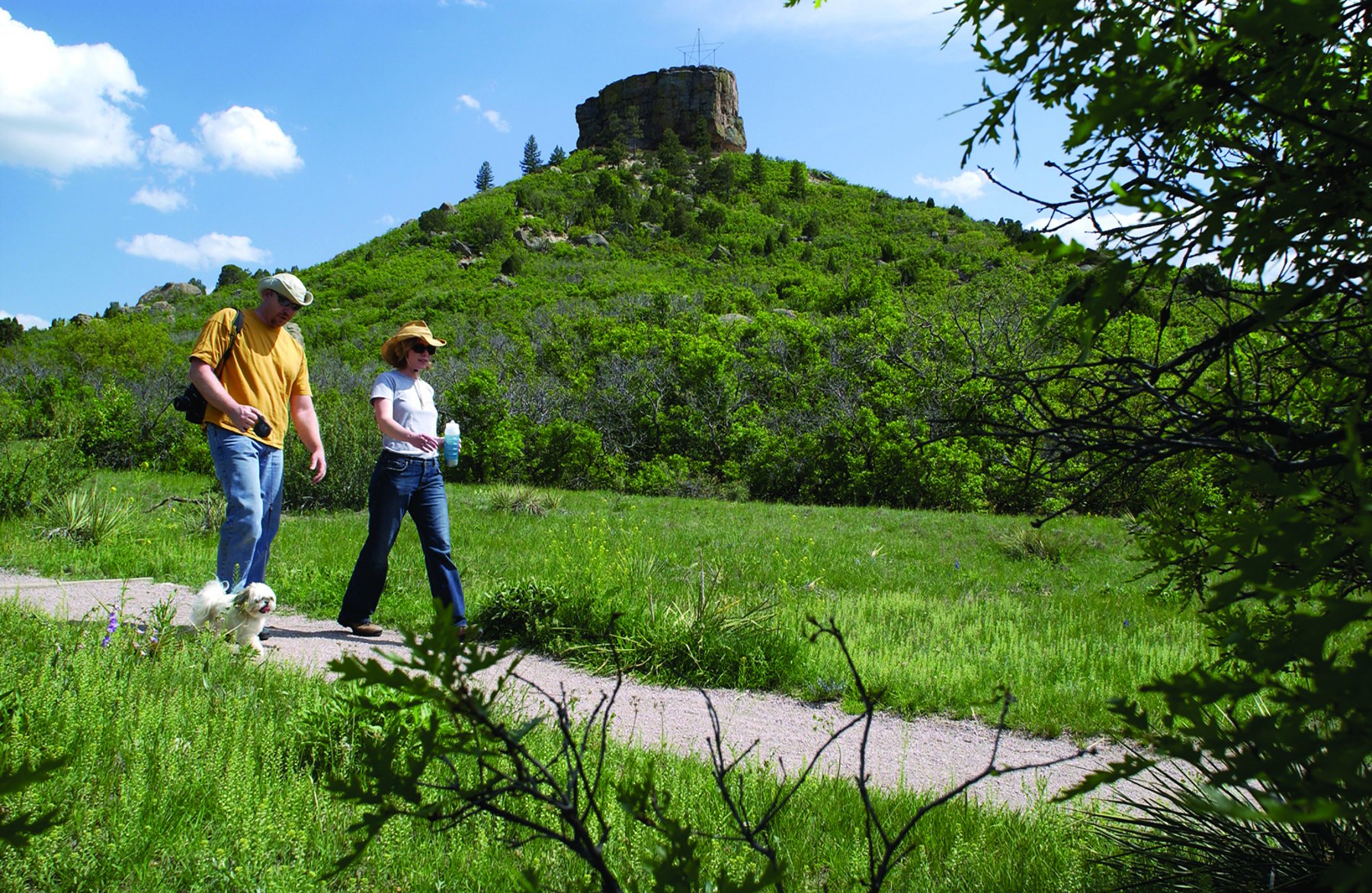 The famous Castle Rock rock sits a top a Kelly-green hill under a bright-blue sky with two people walking a small white dog along a path surrounded by grass.