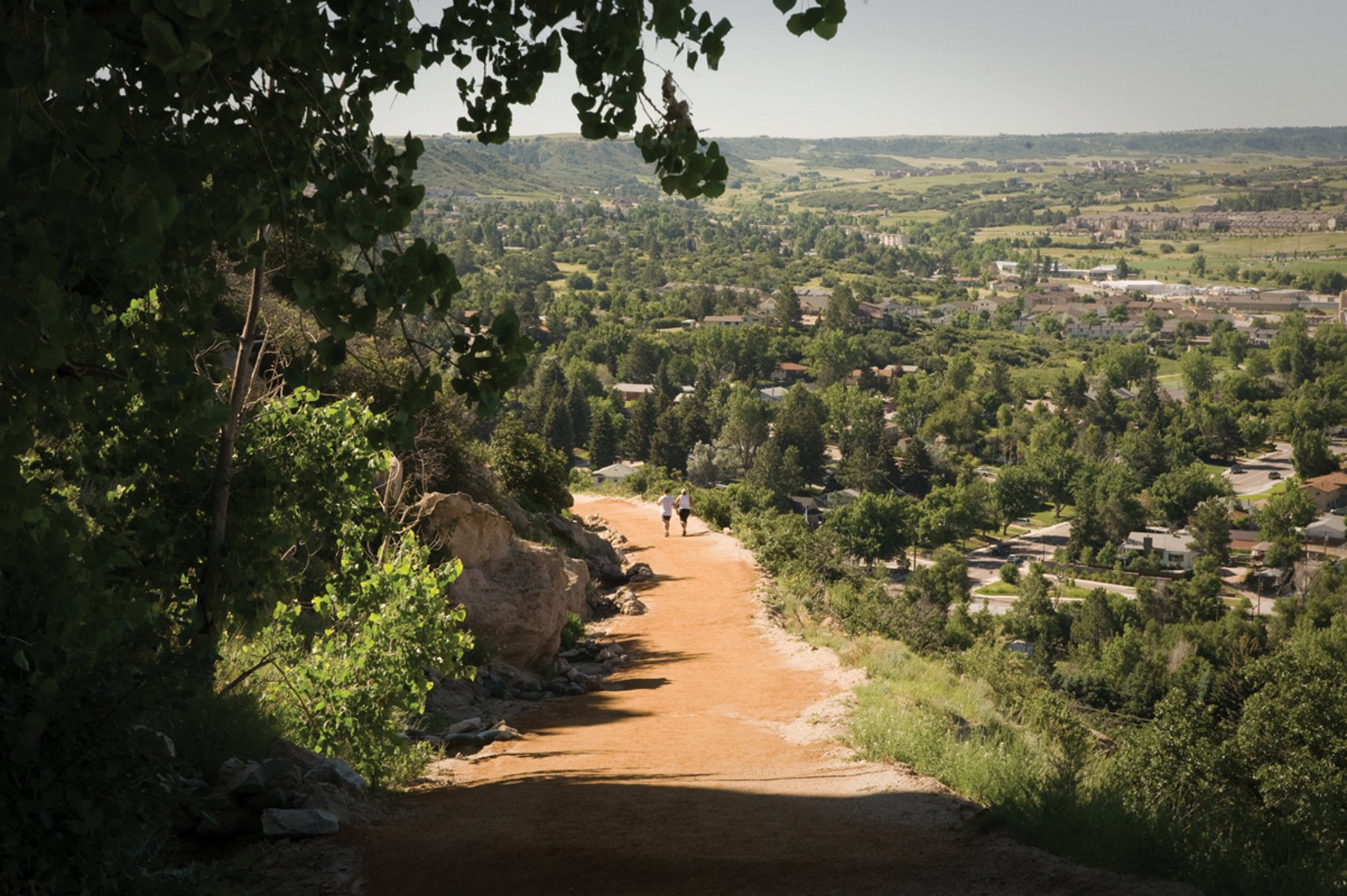 A red-clay path sits on the side of a mountain with greenery on both sides. In the distance, two people walk down. In the background the town of Castle Rock sits. 