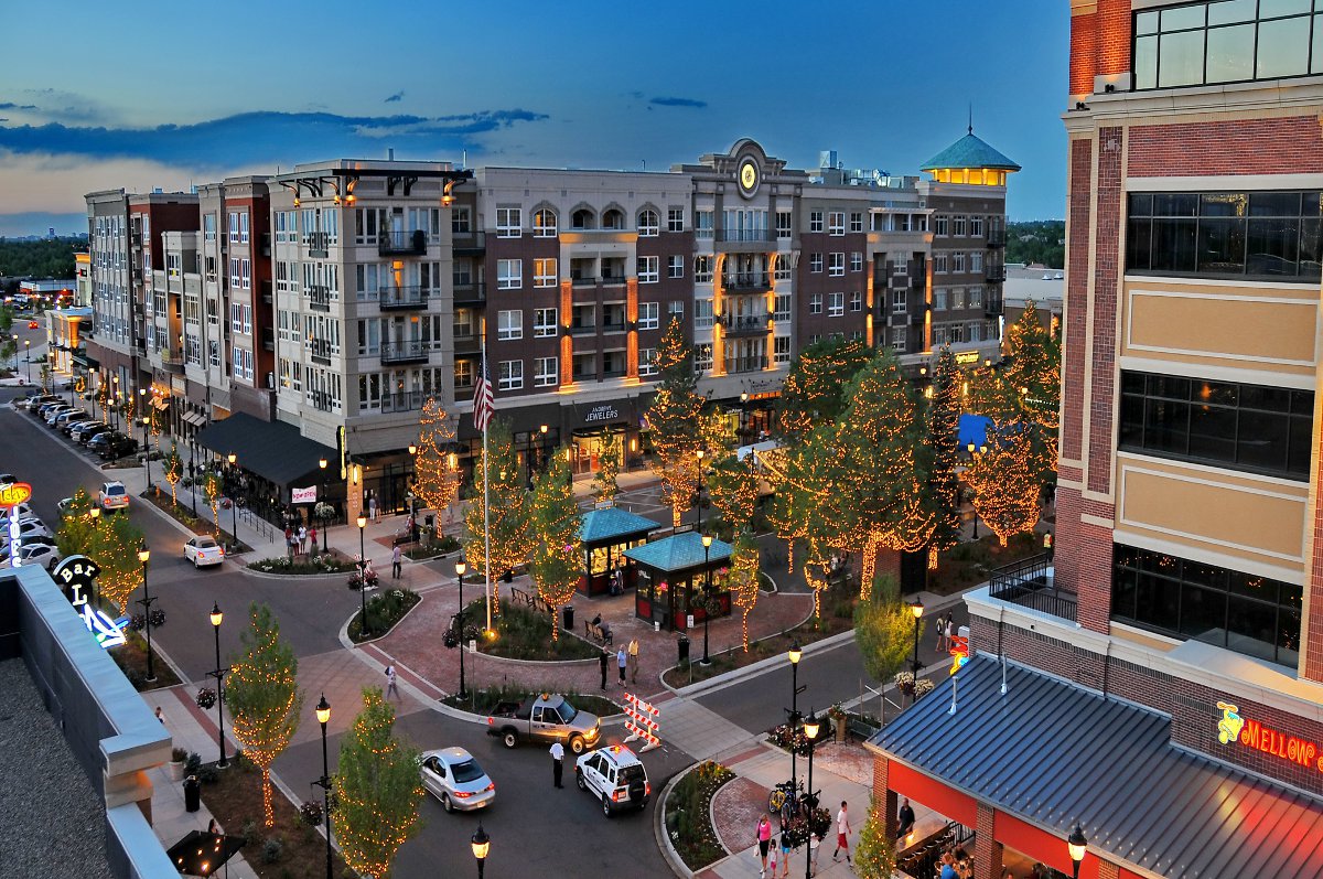 A busy dusk at The Streets at SouthGlenn. People are walking on the sidewalks and cars are in the streets. There are twinkle lights in the trees down the middle of a square and a tall apartment building sits in the middle of the image.
