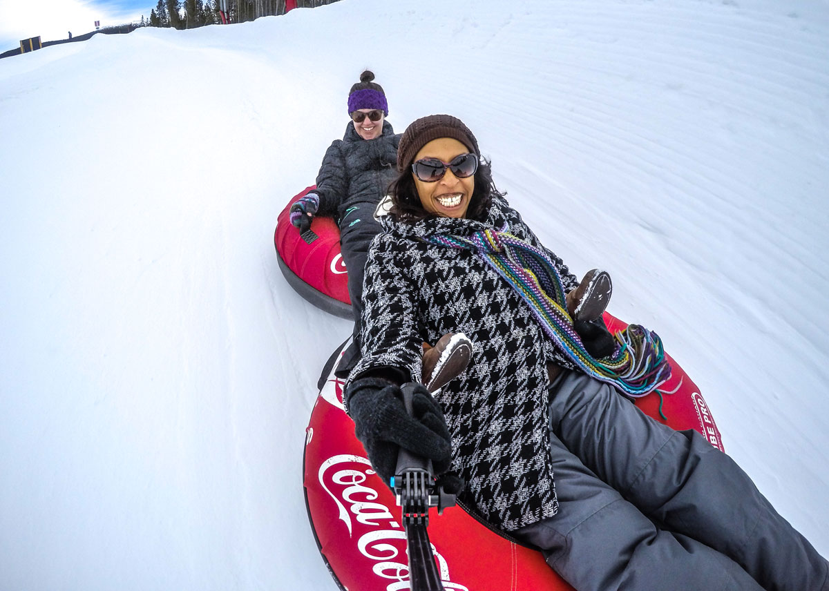Two smiling people dressed warmly slide down a snowy hill in their own red Coca-Cola tubes on the Coca-Cola Tubing Hill in Winter Park. The person in the front is holding a selfie stick as they take a happy photo of them and their friend behind them.