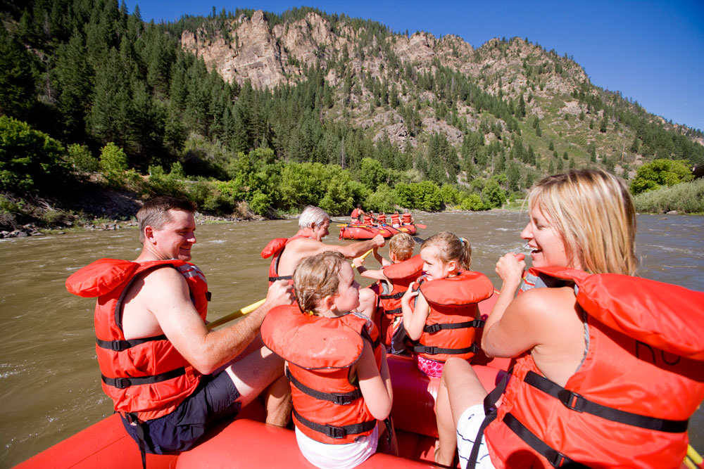 Six people sit in a red inflatable raft wearing red life jackets as the tube down the brown Colorado River with canyon walls covered in evergreen trees.