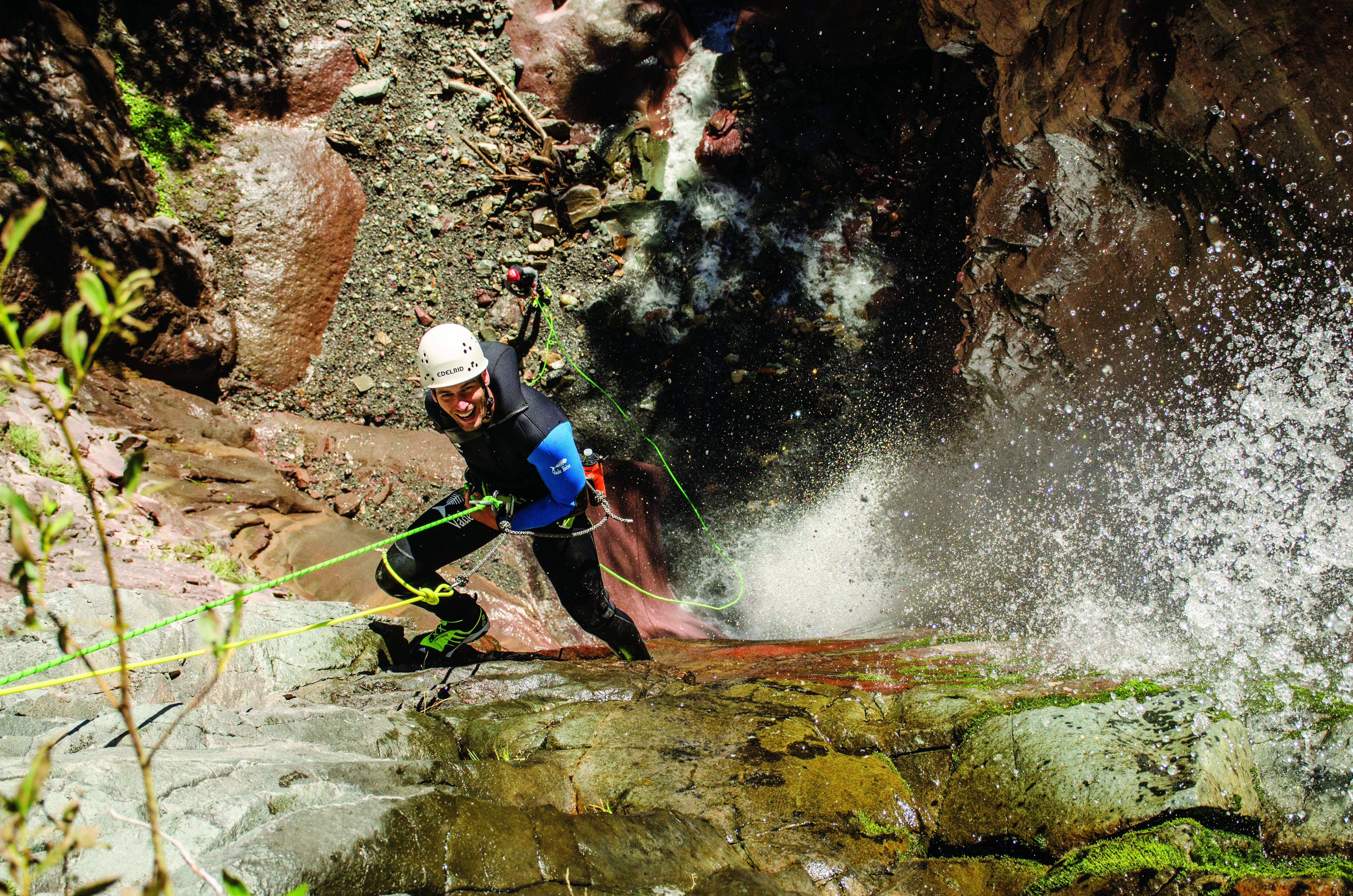 A climber in a wet suit, helmet and climbing harness smiles and looks up as they scale the rocky wall next to a flowing waterfall in Colorado.
