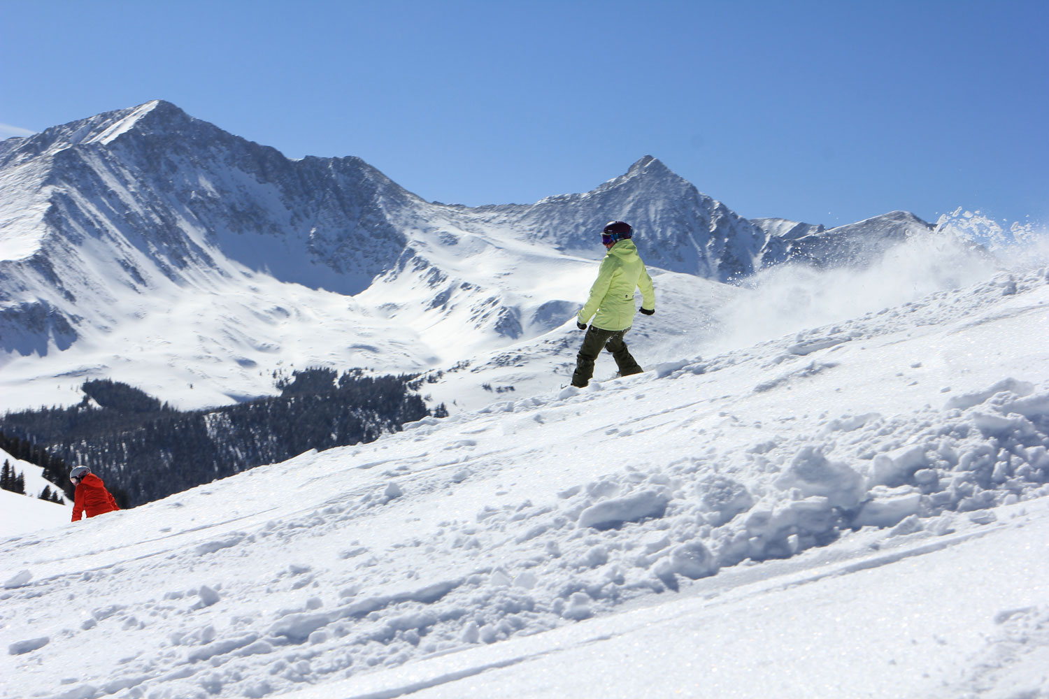 A snowboarder in a green jacket boards down a snowy slope at Copper Mountain with snowy peaks in the distance.