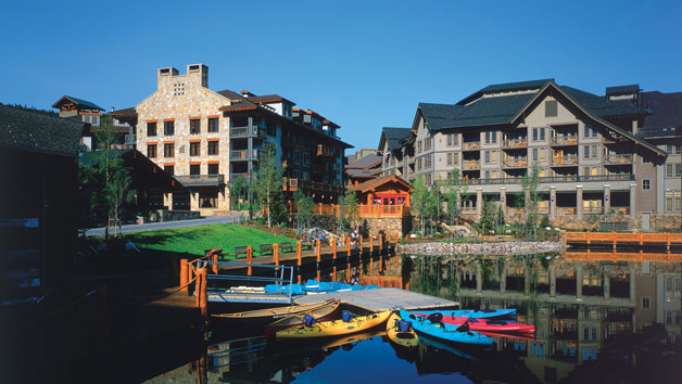 Colorful kayaks sit on a glassy, still lake on a summer's day in Copper Mountain. In the background stone four story buildings sit under a blue sky.