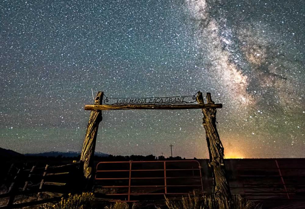 The night sky with the Milky Way behind a wooden gate entrance that says "Luis Maria Baca Grant."