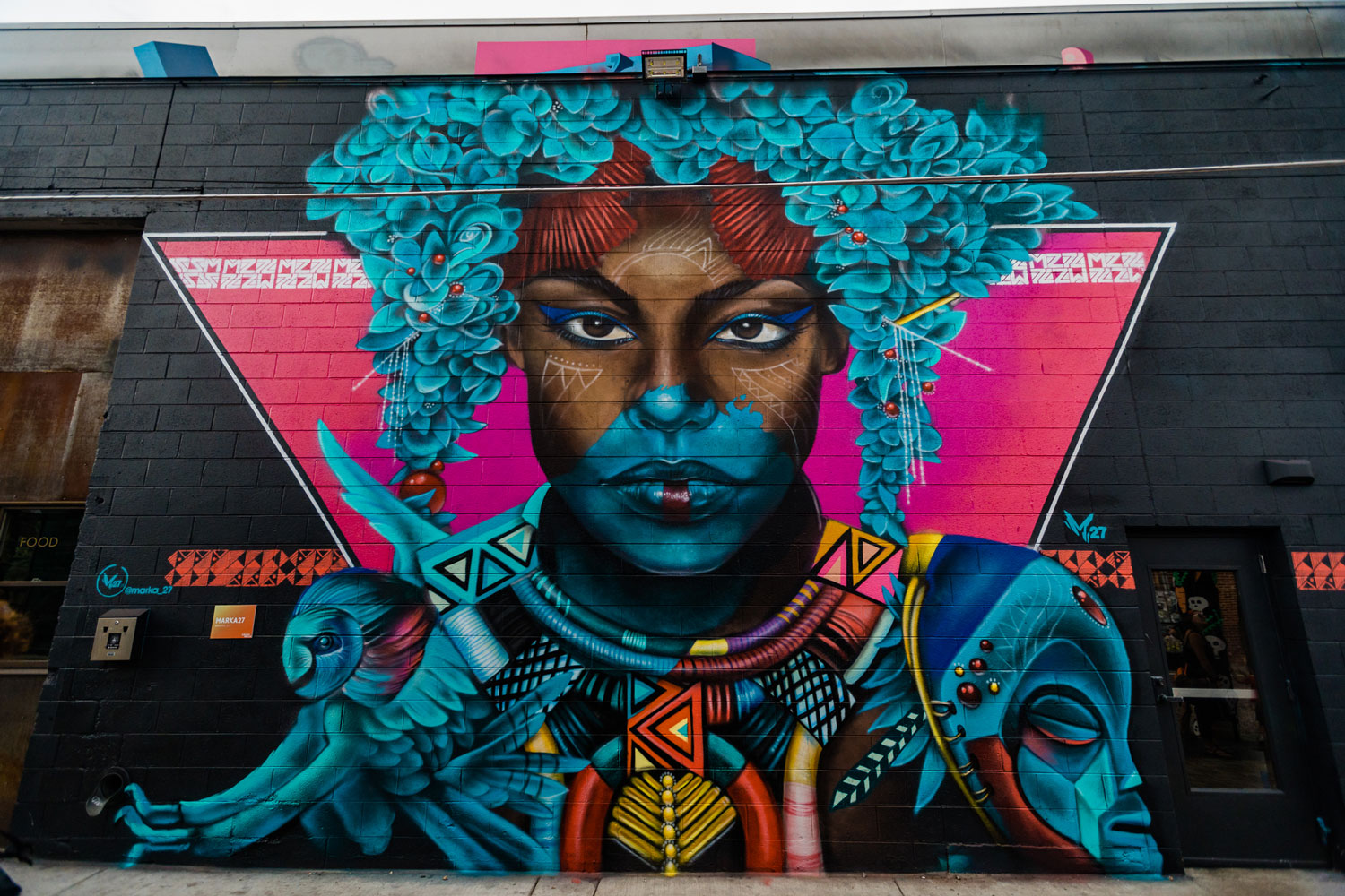 A mural on the side of a black-painted brick building in Denver, Colorado, features an person with vibrant turquoise flowers dripping down from their head.