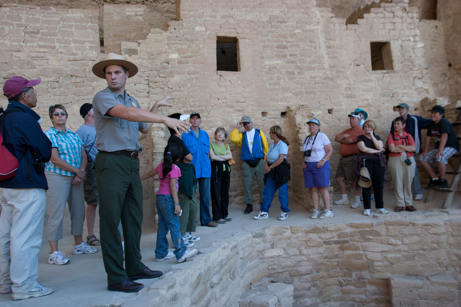 A national park ranger leads a tour of many people standing above a stone cliff dwelling basement with cliff dwelling walls with windows behind the group.