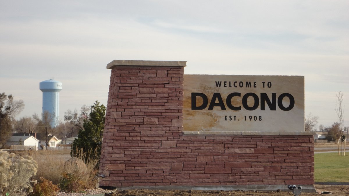 A sign reading "Welcome to Dacono Est. 1908" sits on the right. The portion with writing is on a a solid white stone that sits in the L-shaped base that's made of red, thin, rectangular stones. In the background a water tower sits with houses at its base.