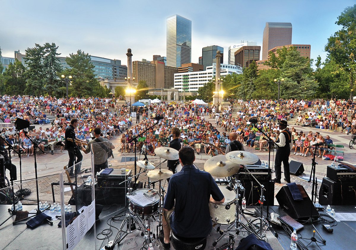A band performs on a raised, outdoor stage for a large audience in Denver, Colorado.