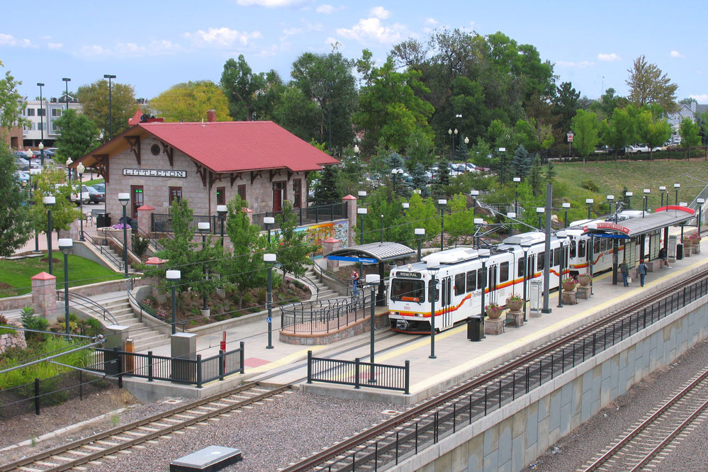On a summer's day the Denver RTD train has pulled into the downtown Littleton station which sits up a set of stairs and is stone with a red roof. Surrounding the tracks are green grasses and green-leafed trees.