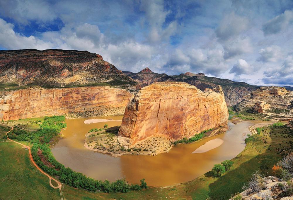 Multi-layered rock plateaus sit in the background under a blue, cloudy sky. In the middle there's a red rock surrounded by the confluence of the Yampa and Green Rivers. It's summer and the riverbanks are covered in green grass and scrubby green trees.