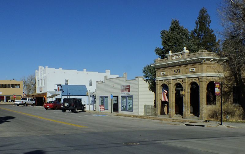 A summer's day in downtown Elizabeth. Sitting on the street, the yellow-brick bank building sits next to a wooden building with a two-story bright-white building farther down. Cars are parked diagonally under a blue sky.