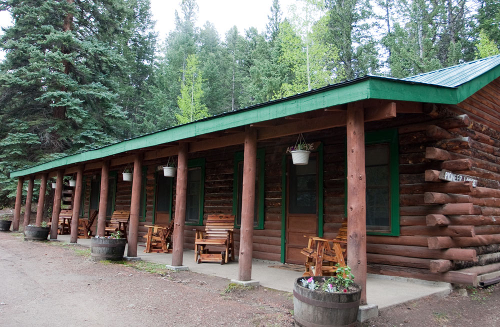 A brown log building features a green roof and green trim around several doors and windows at a ranch near Buena Vista, Colorado. Modern, wooden rocking chairs sit outside each door.
