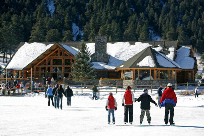 Families and friends skate along an ice rink in Evergreen. A snow-covered lodge sits nearby at the base of a pine-forest-covered mountainside.