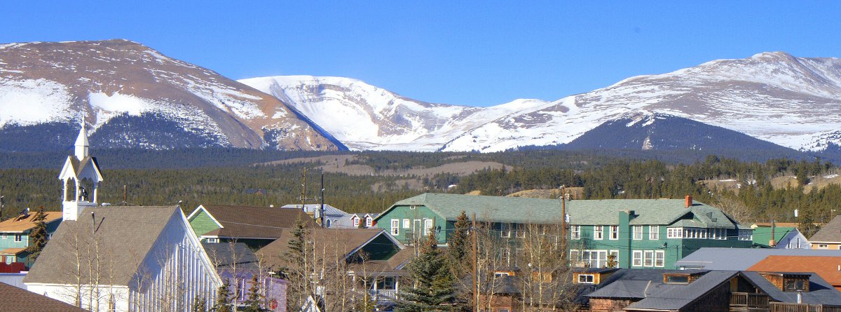 Snow-covered mountains meet a bright-blue sky in the distance behind Fairplay's historic downtown. There are evergreen trees.