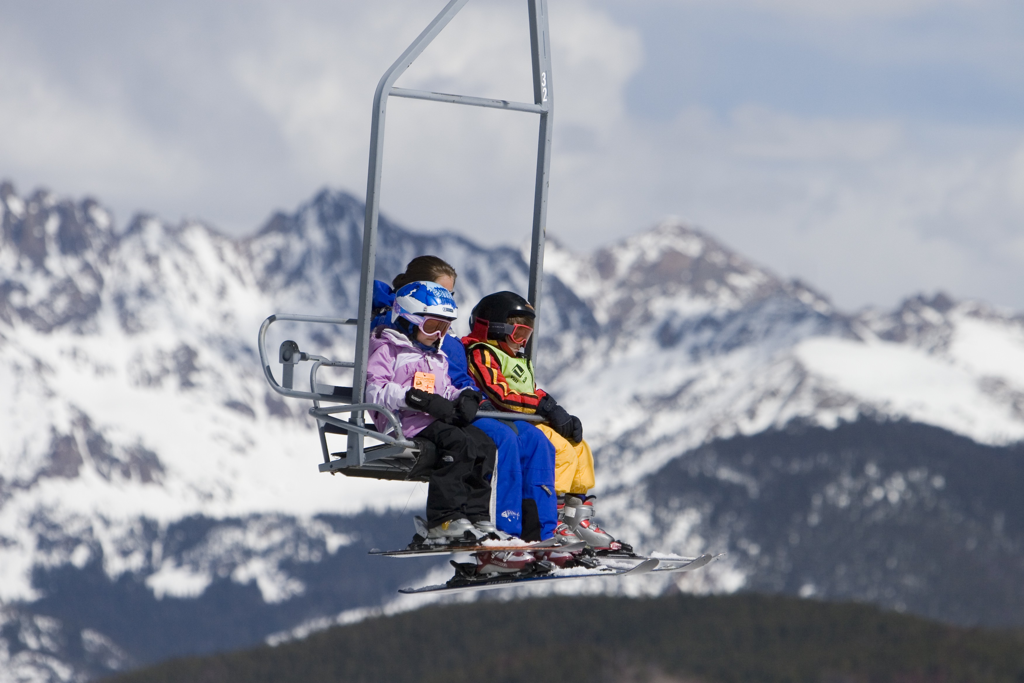 A mom and two kids ride a chairlift with all their ski gear. Behind them, we see snowy peaks.