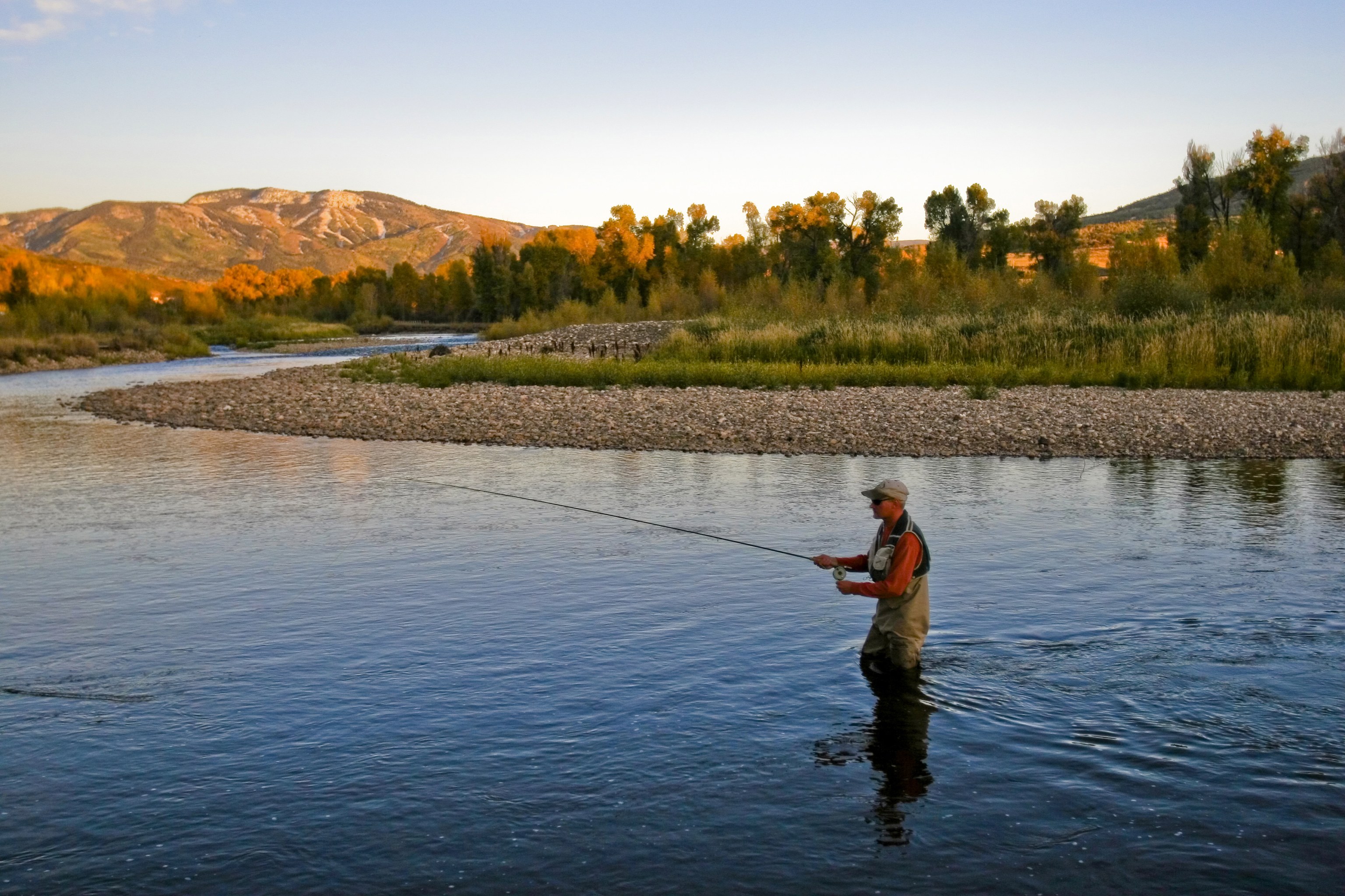A man fly-fishes at dusk in the middle of a calm river. In the background, the mountains are highlighted in golden hues and the trees and grasses along the riverbank are green.