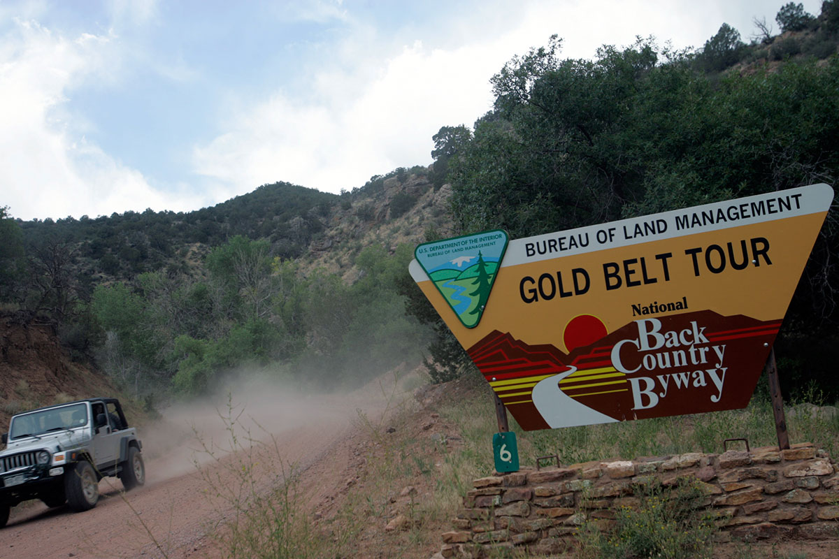 The trapezoid entrance sign reads "Bureau of Land Management Gold Belt Tour National Backcountry Byway." On the left a white jeep drives down a dirt road. It's summer with green trees and a blue-white sky.
