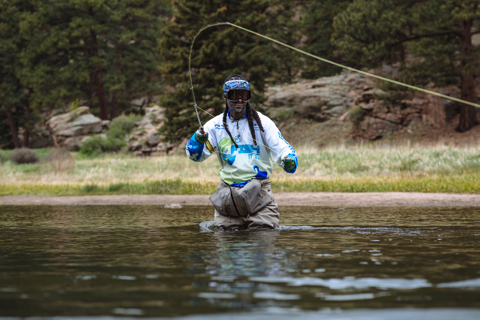 A man stands waist deep in a river, casting his rod and reel