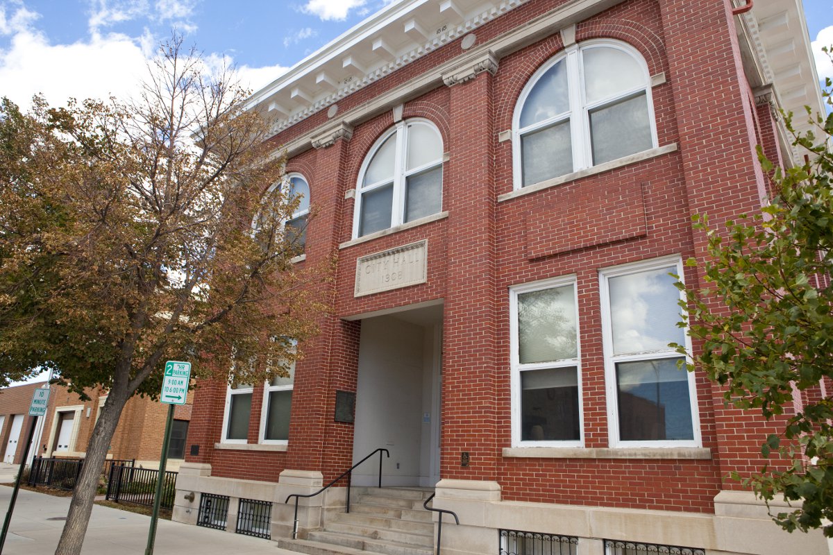 A two-story red-brick building sits at an angle with rectangular windows on the first floor and arched windows on the second floor. There are green trees at the street level.