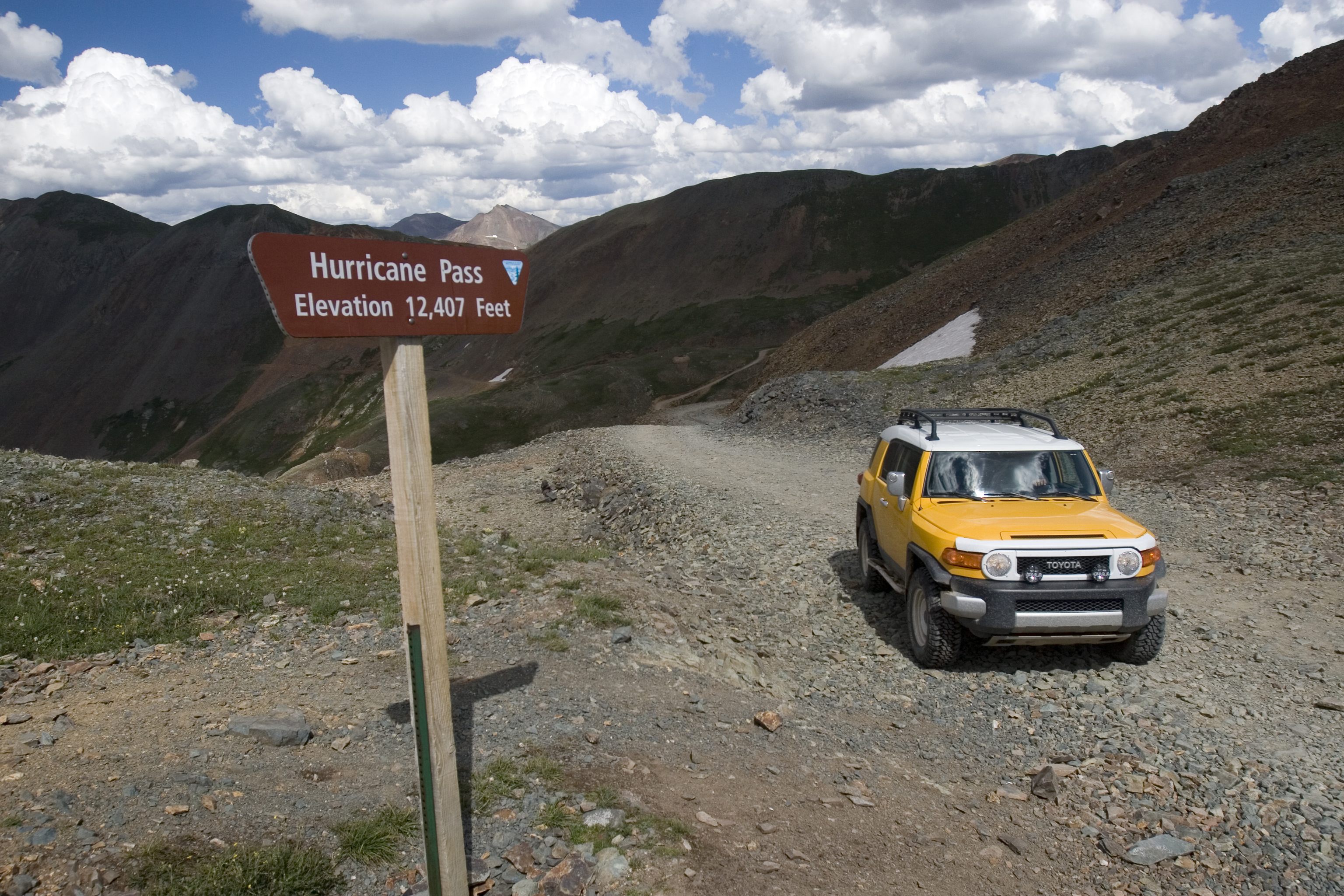 A canary yellow all-wheel-drive vehicle climbs a dirt road. A brown forest service sign reads "Hurricane pass. Elevation 12,407 feet"