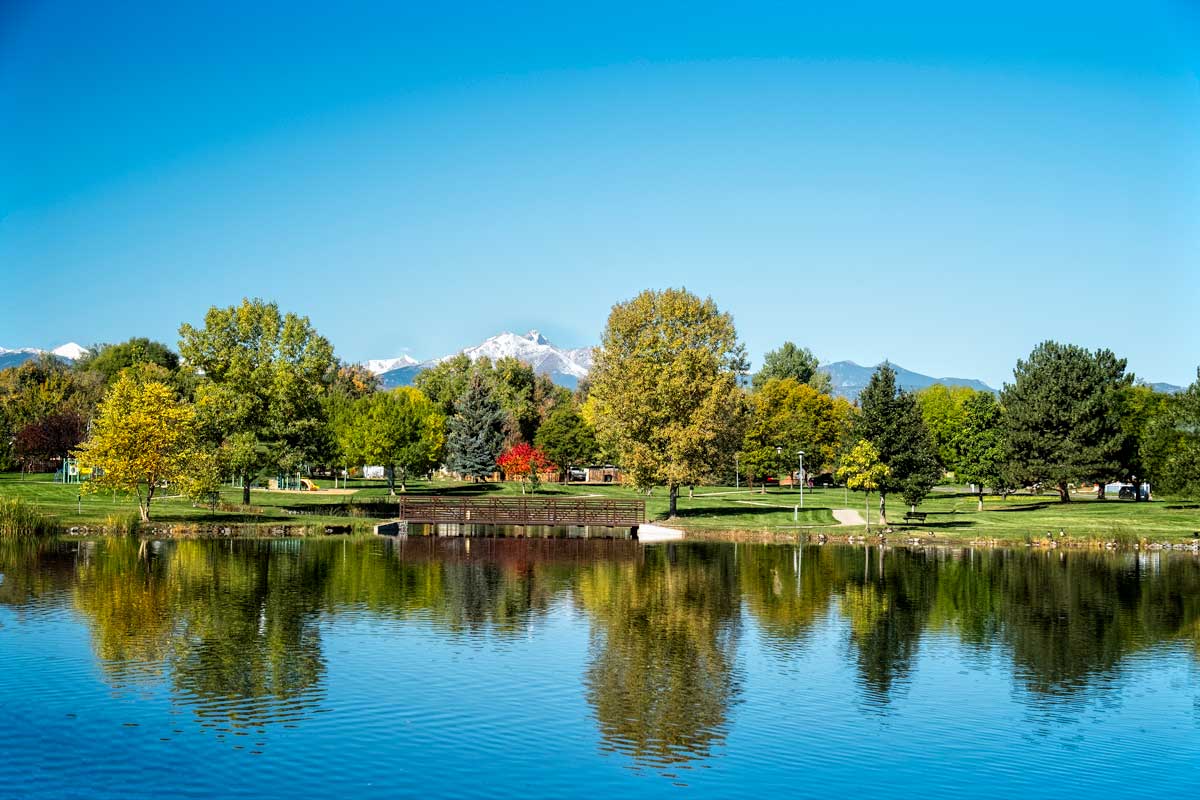 A calm lake reflects green-leafed trees, a pedestrian bridge and pathways at Loomiller Park in Longmont. In the distance a snow-capped mountain sits beneath a blue sky. 