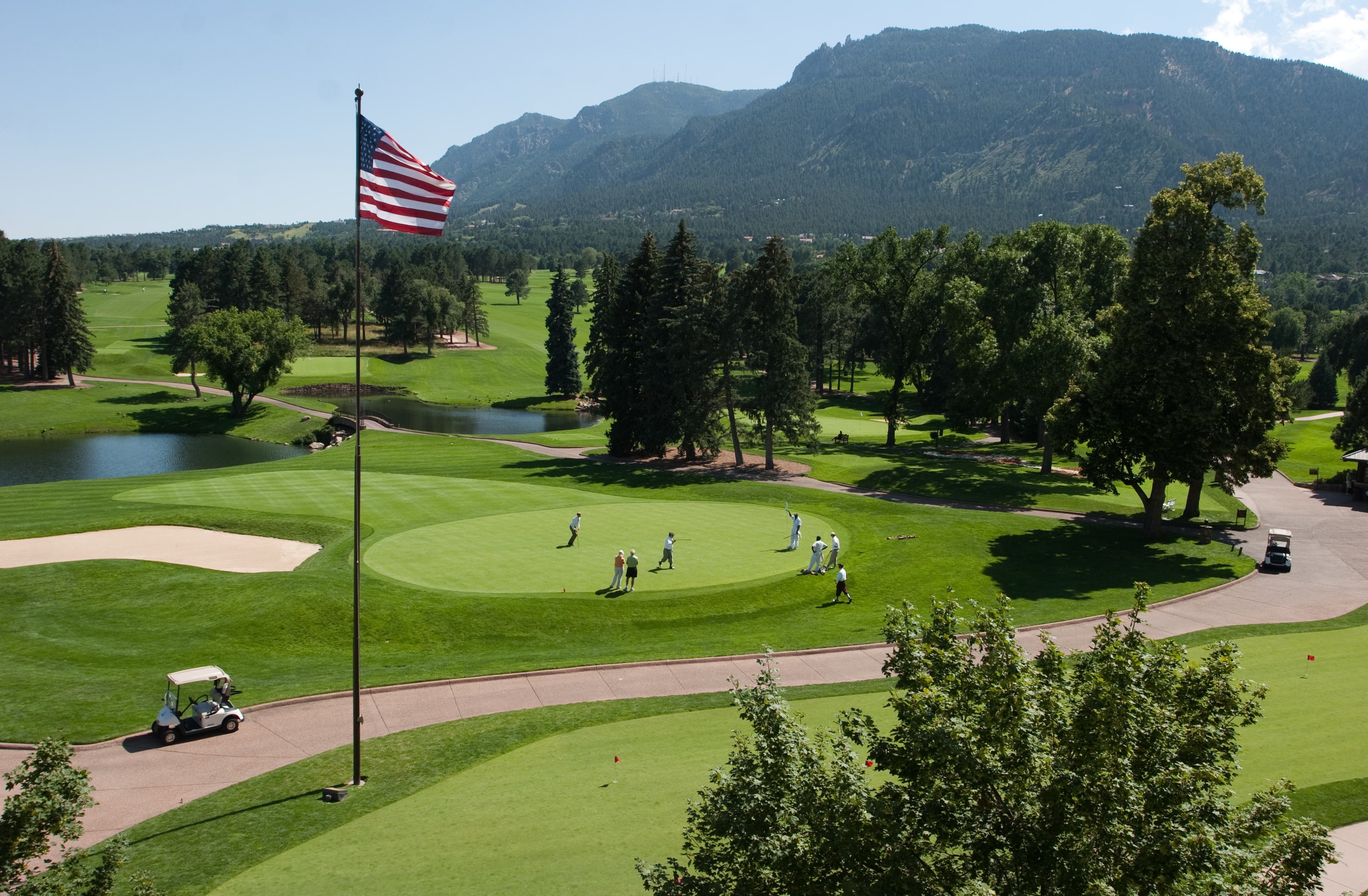 An American flag waves over the bight-green course. From a distance, we see tiny golfers making their puts. Two golf cards ride a red-brick road, and a forested hill looms in the distance
