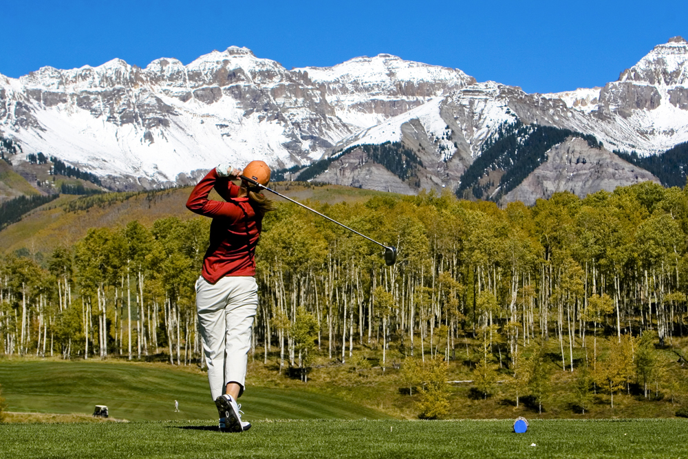 A woman in a bright red top swings for the mountain tops — literally! We can see the jaggest snowcapped peaks of Telluride in the distance. Beneath those, a stand of aspen trees is starting to changes to fall's yellow leaves.