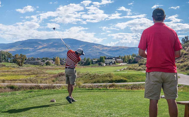 A man swings his golf club with another man in a pink shirt looking on. The green grass leads to houses in the distance and in the hazy background mountains rise up.