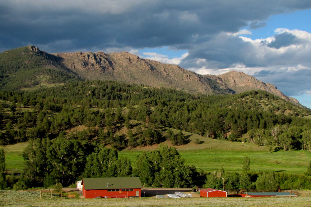 Two red barns sit at the base of rolling green hills covered in green trees beneath rocky, grey mountains underneath a blue sky with grey clouds.