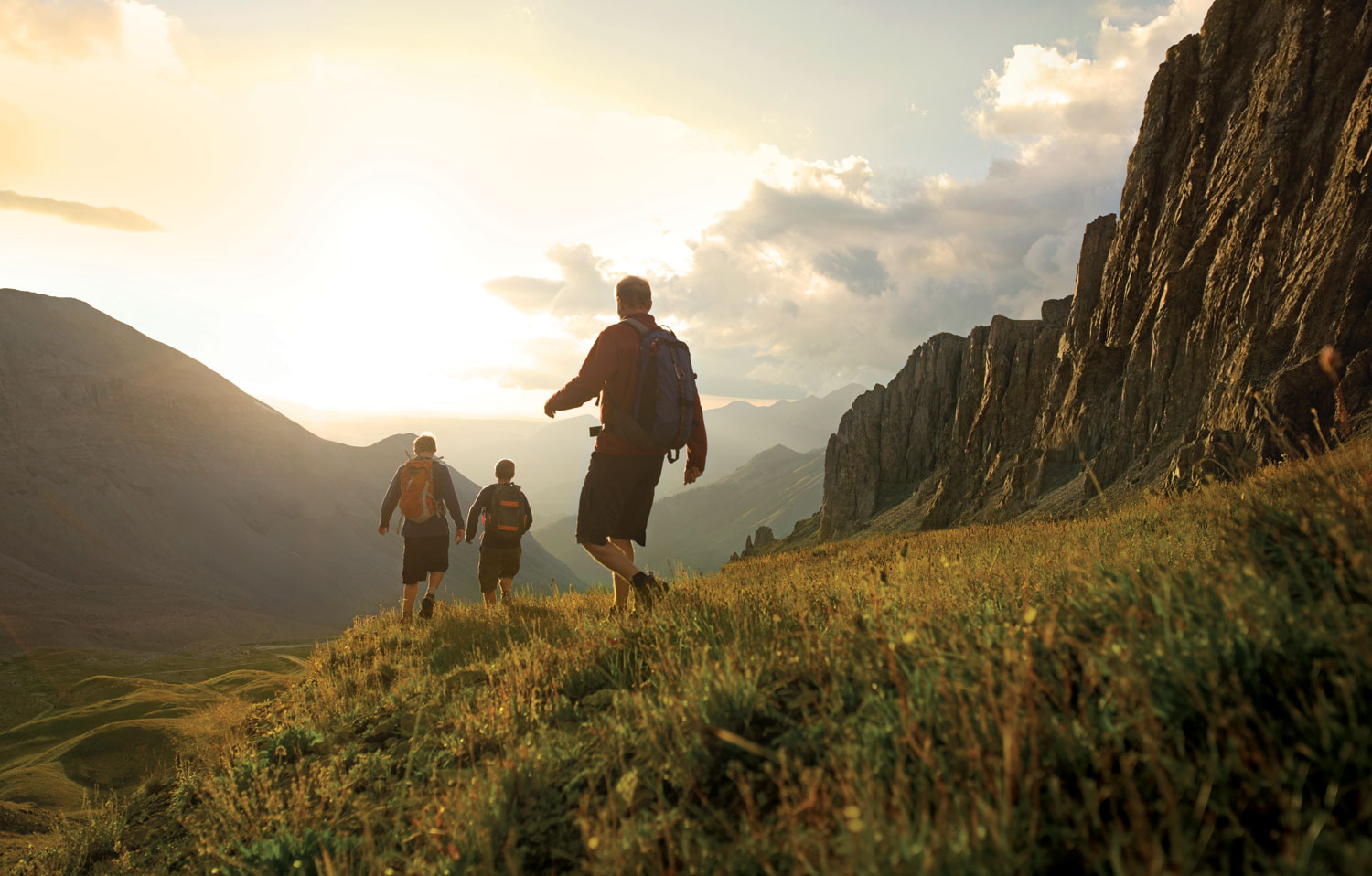 Three hikers head downhill between to rising peaks, toward the sun, which is setting