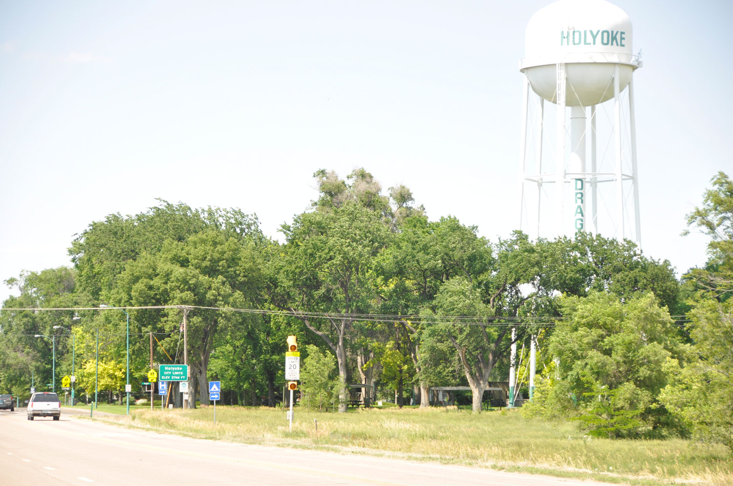 A white water tower that says "Holyoke" sits over green trees and a road with two cars on it.