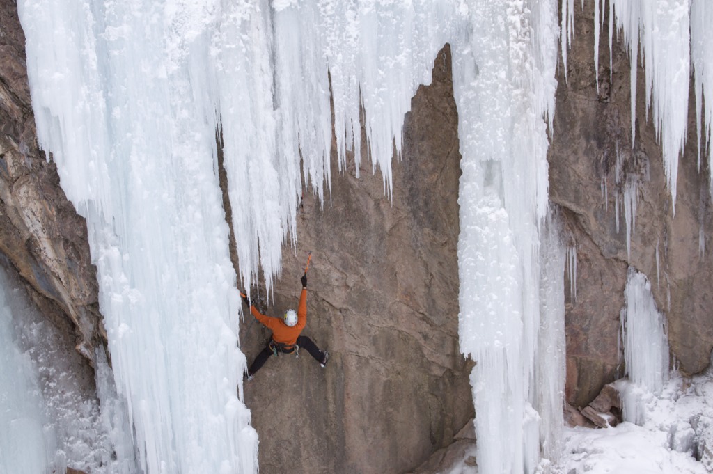 A climber in a burnt-orange jacket and silver helmet makes their way up a rock wall covered in ice near Ouray, Colorado.