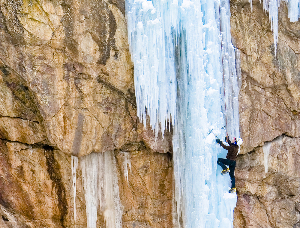 A climber in black scales a section of ice wall using ice picks and spiked boots near Ouray, Colorado.
