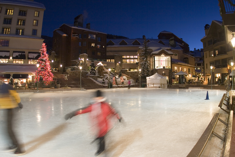 Blurry ice skaters skate around a rink surrounded by Beaver Creek Village buildings at dusk. 