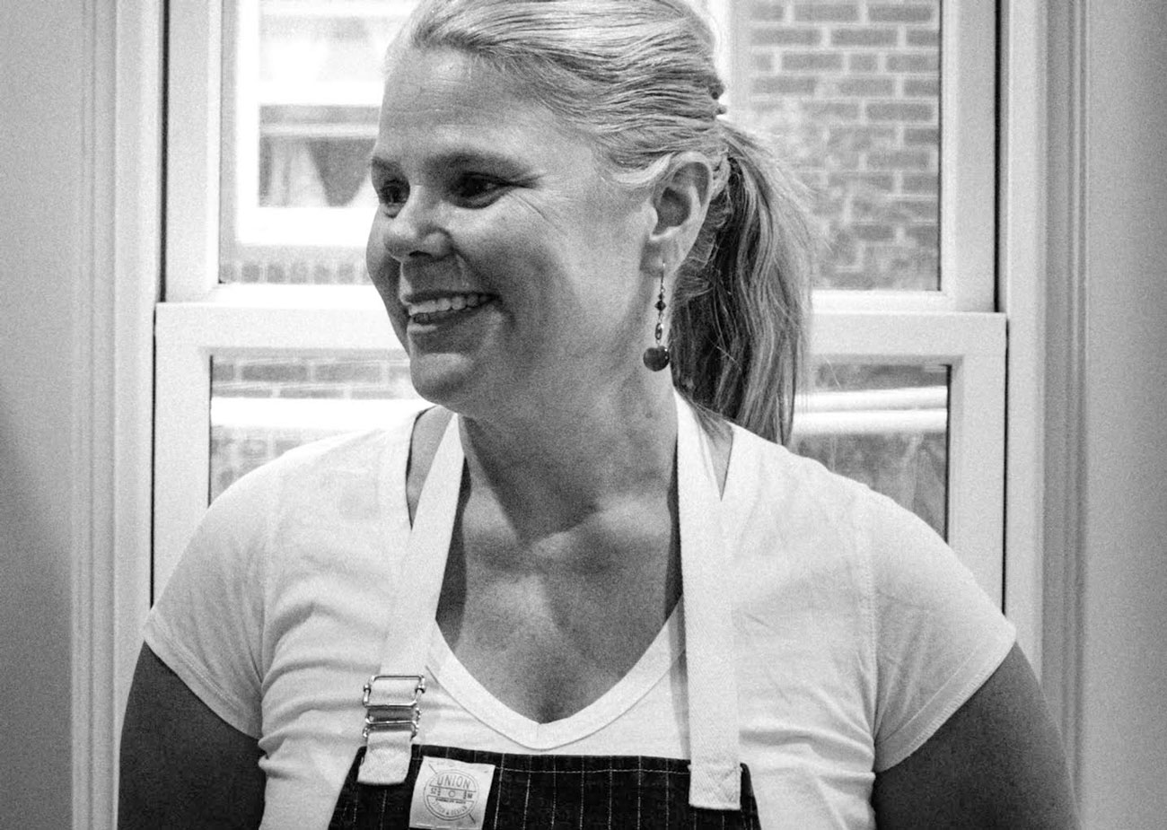 Shown in balck and white, chef Jennifer Jasinski, of Denver's Rioja, Stoic & Genuine and Bistro Vendôme, wears a cooking apron and smiles at someone out of view.
