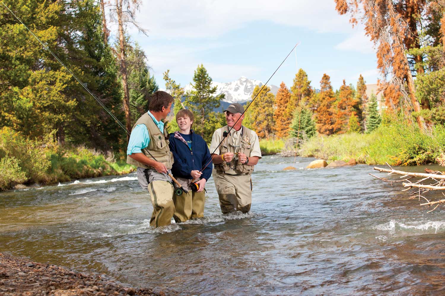 Two men and a teen in fishing waders hold fly-fishing poles and stand knee-deep in a river near Keystone, Colorado.