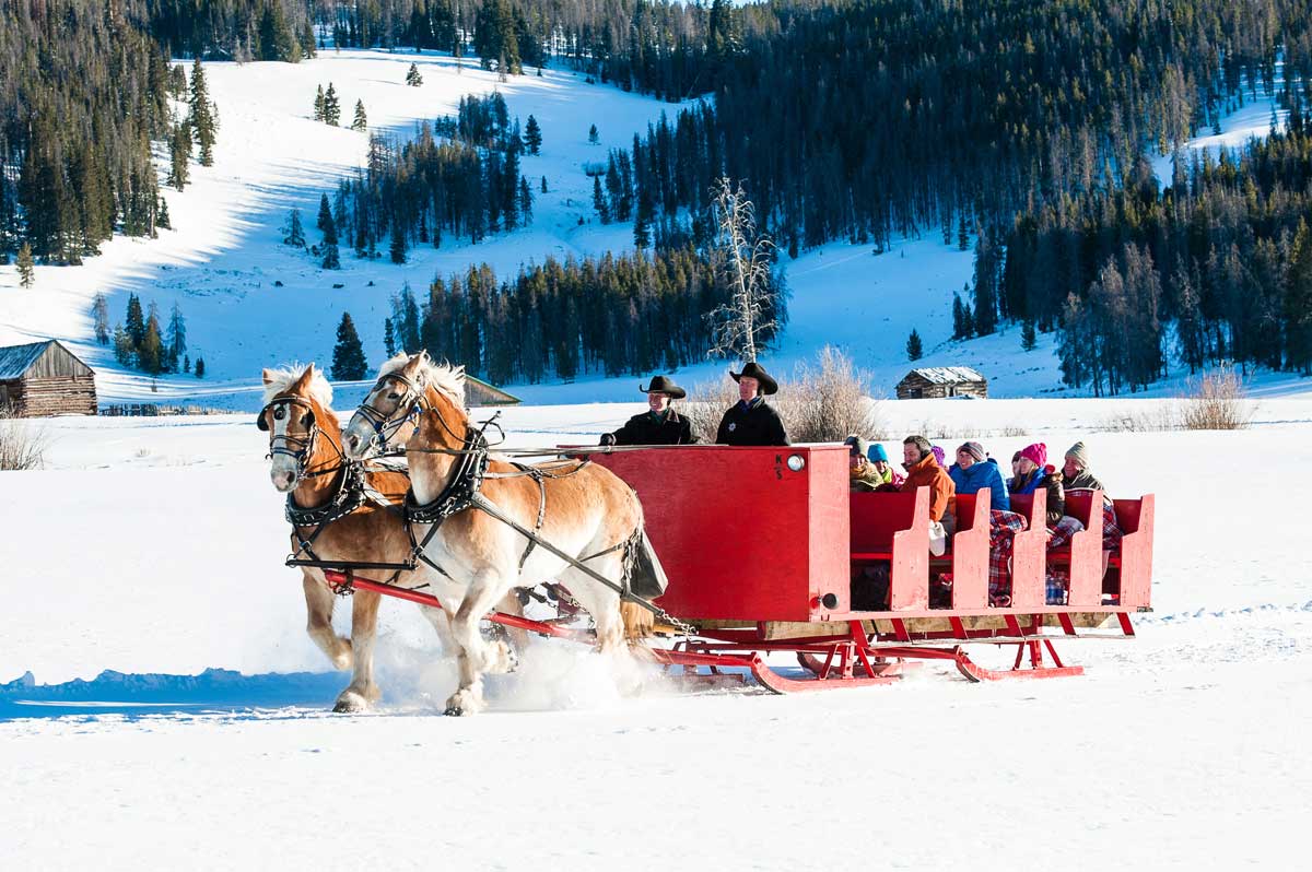 Two massive matching horses pull a red sleigh with two drivers and four rows of passengers through a snowy field at Keystone Resort in Colorado.