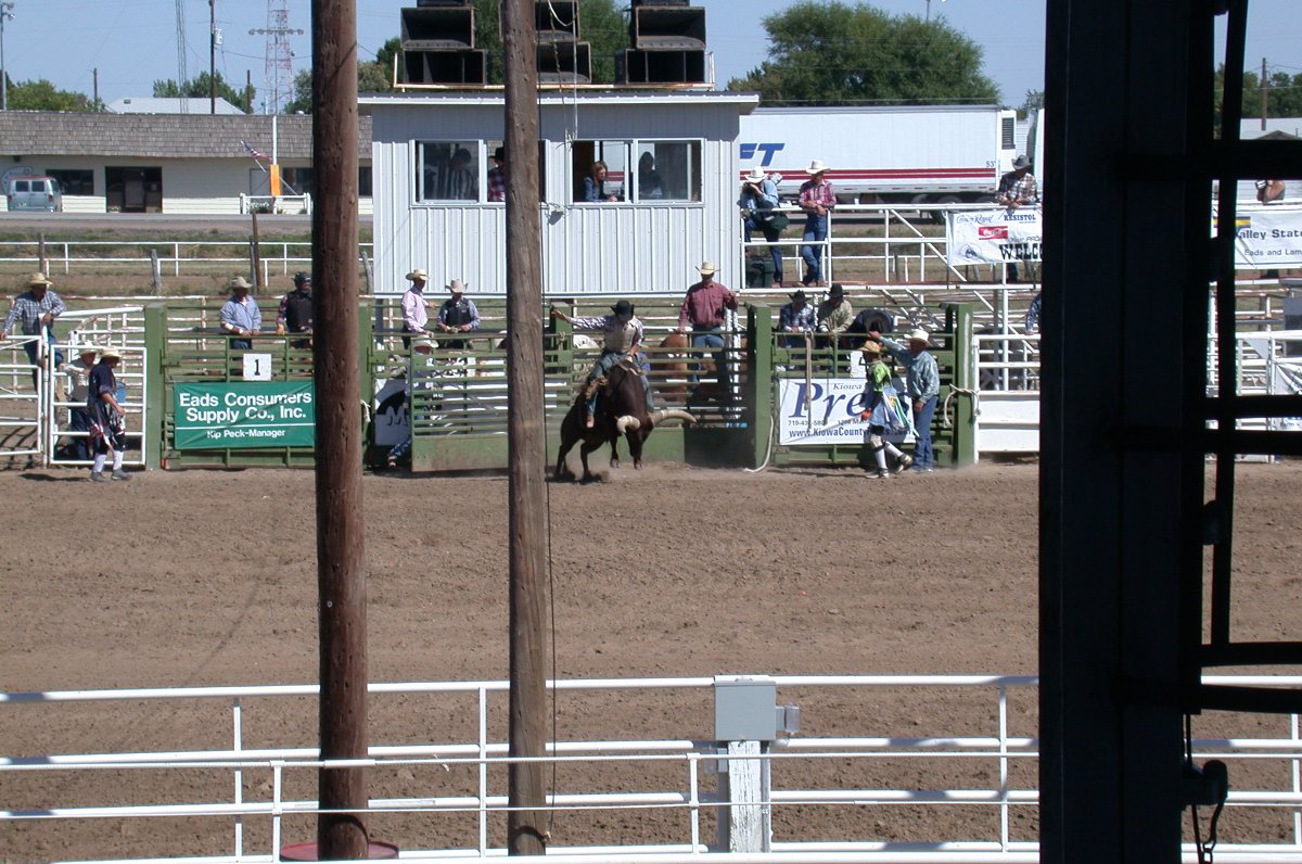 Through the the stands, a bull rider leaves the paddock. There are other people behind in the paddocks and to the right and left of the fences at a rodeo in Kiowa. 