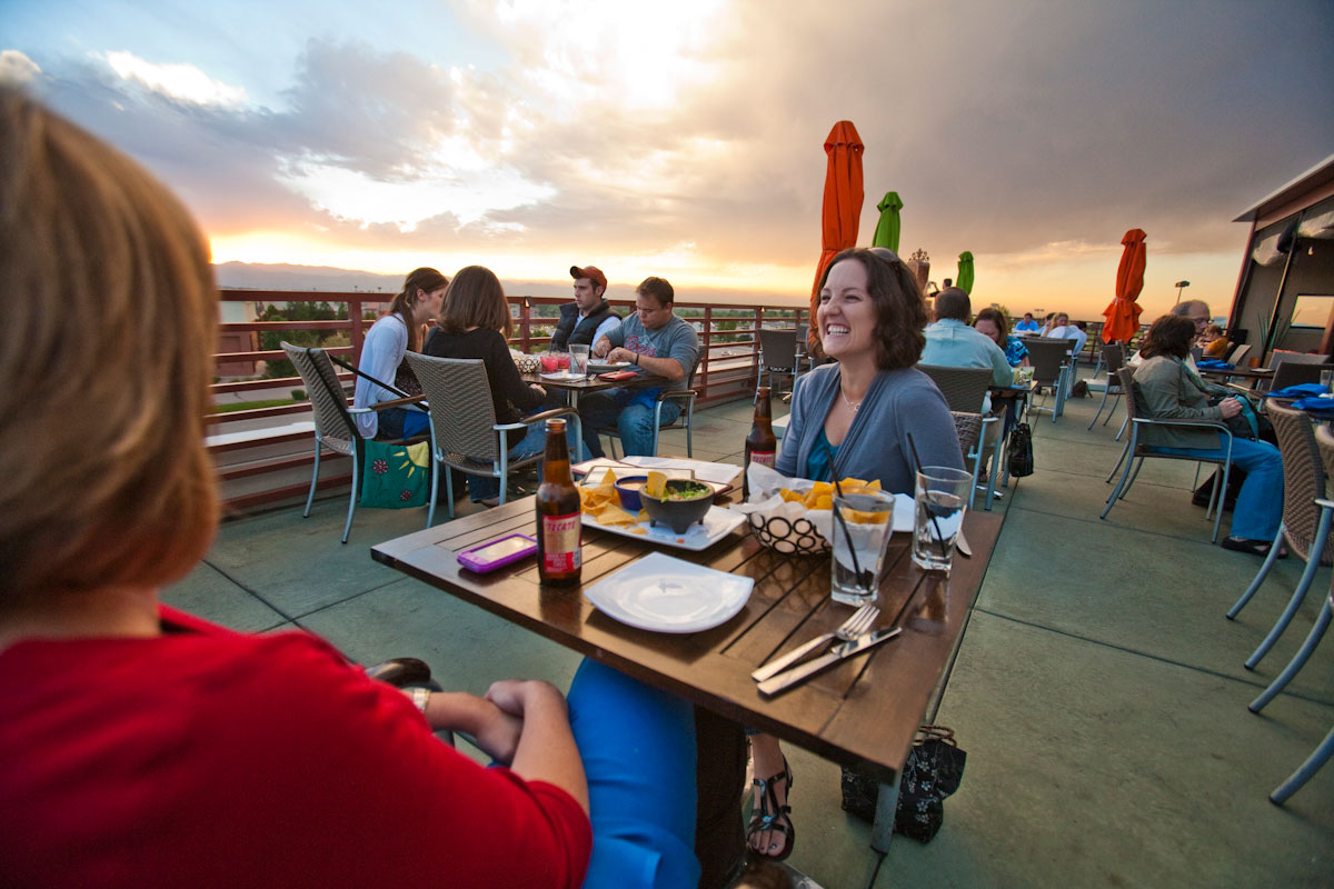 People sit on a rooftop eating dinner with a pink, yellow and blue sky up above.