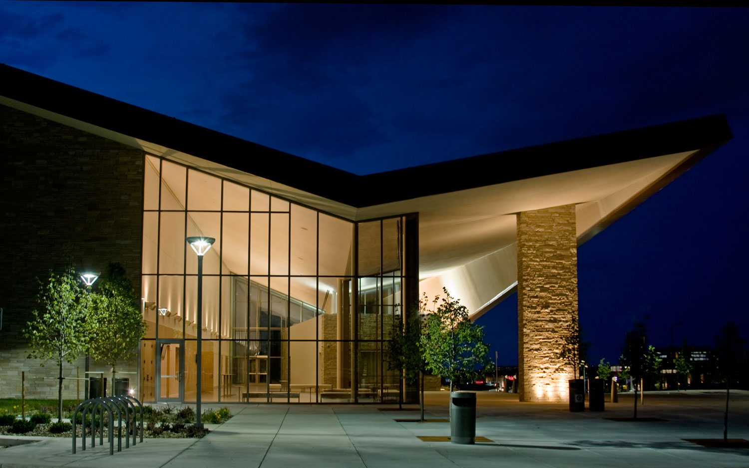 A building with a angled roof sits lit up at night with young green-leafed trees, trash cans and bike racks out front.