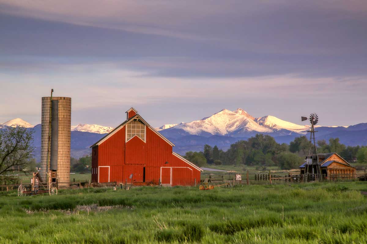 A red barn with a grey silo and a brown wooden house sit in a green-grass field with views of snow-capped mountains in the distance. 