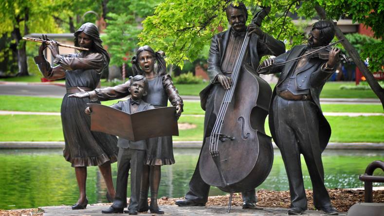 A piece of public art made of metal with a woman playing the flute, two children singing, a man playing a base and a man playing a violin near a lake.