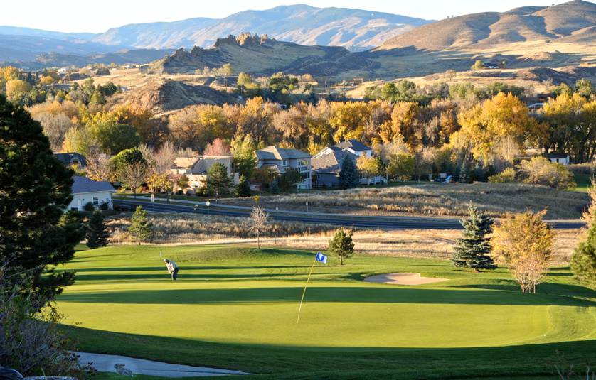 The green of a golf course is bathed in late afternoon sunlight as a lone golfer waits to strike their ball. In the background houses are among trees with fall leaves and the foothills are rolling.